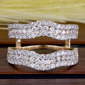 May include: A gold-toned ring guard with two curved bands encrusted with small, sparkling diamonds. The ring guard is designed to sit around a center stone. The background is a blurred mix of yellow and brown.