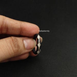 May include: A black ring with a silver band featuring three diamond-shaped accents. The ring is held in a hand against a dark background. The text "Bellaeternity" is visible.