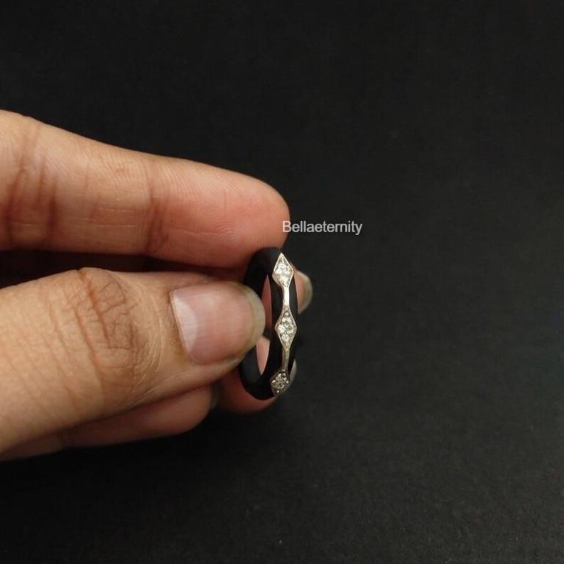 May include: A black ring with a silver band featuring three diamond-shaped accents. The ring is held in a hand against a dark background. The text "Bellaeternity" is visible.
