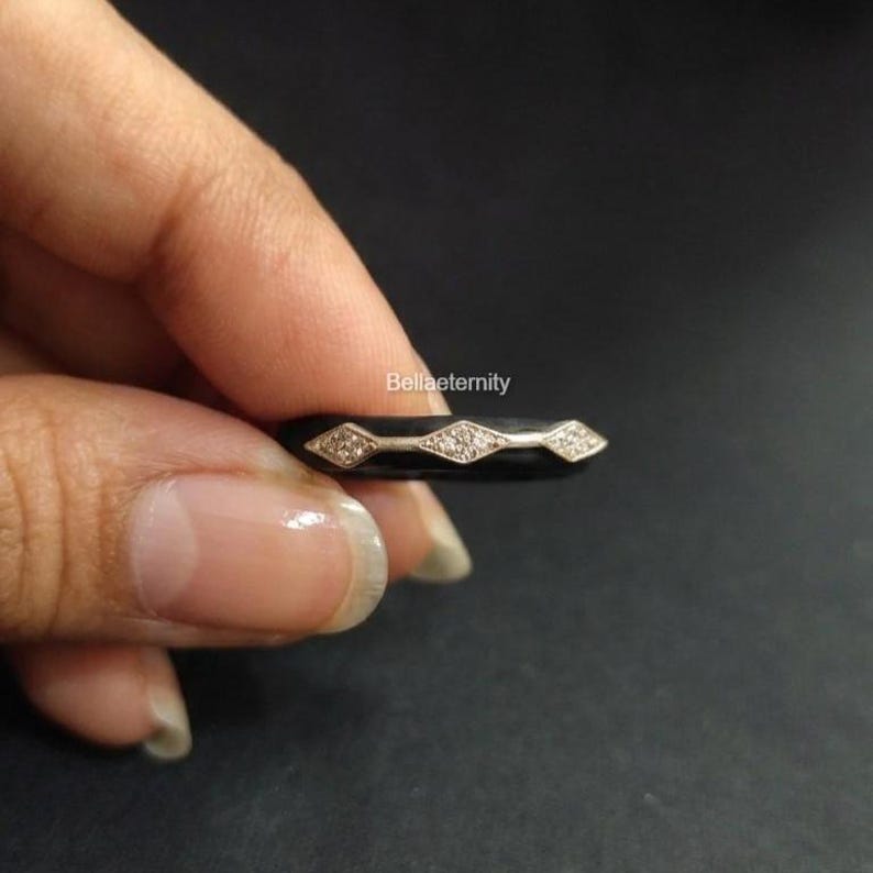 May include: A black ring with three diamond-shaped sections, each with small, sparkling stones. The ring is held against a dark background, showcasing the contrast of the shiny stones against the black band. The ring is a piece of jewelry.