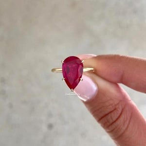 Pode incluir: Um anel de ouro com uma grande pedra preciosa vermelha escura em forma de pêra. A pedra preciosa é presa por quatro garras. O anel tem uma banda fina. O anel é segurado por uma mão.
