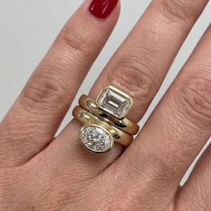 Puede incluir: Tres anillos de oro con piedras preciosas transparentes. El anillo superior presenta una piedra de talla esmeralda, el anillo del medio es una banda simple y el anillo inferior tiene una piedra de talla ovalada. Los anillos están apilados en un dedo.