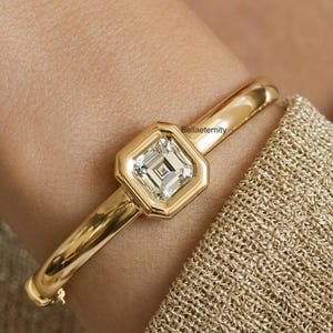 May include: A gold bangle bracelet with a square-cut diamond center stone. The diamond is set in a geometric bezel. The bracelet is a polished gold color and has a clasp closure. The text "Bellaeternity" is visible.