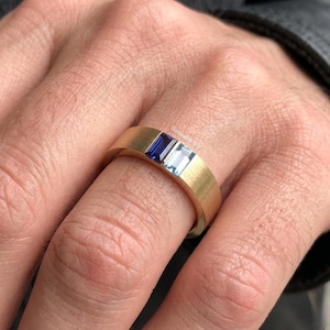 Peut inclure: Une bague dorée avec une finition brossée, ornée de deux pierres rectangulaires : un saphir bleu foncé et une aigue-marine bleu clair. La bague est présentée sur un doigt, mettant en valeur les couleurs des pierres et le design.