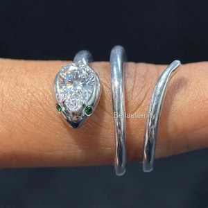 Könnte beinhalten: Silberfarbener Schlangenring mit einem birnenförmigen, klaren Edelstein-Kopf und zwei kleinen grünen Edelsteinen als Augen. Der Ring wickelt sich spiralförmig um den Finger.