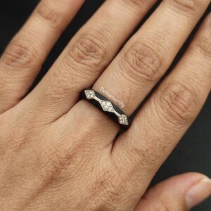 May include: A black ring with a geometric design featuring three diamond-shaped accents. The ring has a silver band and is worn on a finger. The text "Bellaeternity" is visible on the ring.