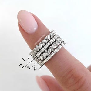 May include: Four silver rings with different gemstone cuts, including baguette and marquise shapes, are stacked on a finger. The rings are set with clear gemstones. The brand name "Bellaeternity" is visible on the finger.