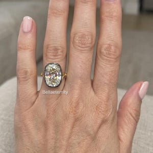 May include: A gold ring featuring a large, oval-cut, light yellow gemstone set in a dark, metallic bezel. The ring band is thin and gold, and the text Bellaeternity is visible below the setting on the finger.