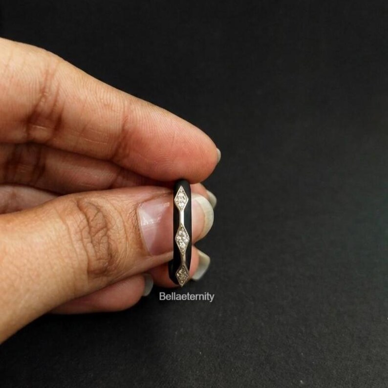May include: A black ring with three diamond-shaped sections, each containing small, sparkling stones. The ring is held against a dark background, showcasing its elegant design. The text "Bellaeternity" is visible on the ring.