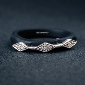 May include: A black ring with a silver band featuring three diamond-shaped sections, each with small, clear stones. The ring has the text "Bellaeternity" on the band. The ring is set against a dark background.