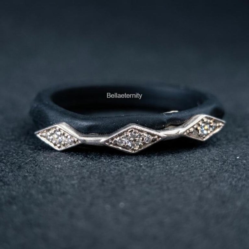 May include: A black ring with a silver band featuring three diamond-shaped sections, each with small, clear stones. The ring has the text "Bellaeternity" on the band. The ring is set against a dark background.