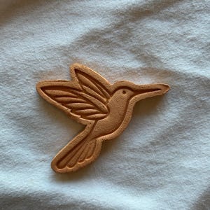May include: A terracotta-colored hummingbird-shaped object. The bird is depicted in profile with detailed wing and tail feather carvings. The object is set against a white, textured background.
