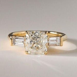 May include: A gold ring featuring a large, central cushion-cut diamond held by four prongs. The center stone is flanked on each side by a single, rectangular baguette-cut diamond. The band is crafted from polished yellow gold with a smooth finish.