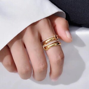 May include: Gold rings in a layered design. One ring has a row of small, clear stones, the others have a smooth, polished finish. The rings are stacked on a finger, creating a modern and elegant look.