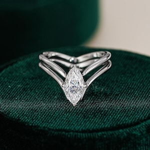 Könnte beinhalten: Ein Verlobungsring mit Diamanten im Marquise-Schliff und geteiltem Schaft-Design. Der Ring verfügt über einen großen, klaren Diamanten, der in einem weißen Metallband gefasst ist und einen modernen und eleganten Stil zeigt. Der Ring wird auf einer dunkelgrünen Samtoberfläche präsentiert.
