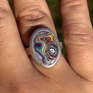 May include: Silver ring with an oval, patterned stone. The stone displays a vibrant mix of colours including blue, green, yellow, and red. The ring's setting has a textured, scalloped edge, adding detail to the design.