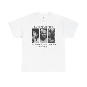 Puede incluir: Camiseta blanca con un diseño gráfico. Presenta retratos en blanco y negro de Michael Jackson, Mike Tyson y Michael Jordan. El texto "TAKE YOUR PICK" y "JACKSON, TYSON, JORDAN GAME 6." están impresos debajo.