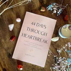 Puede incluir: Un libro rosa titulado "44 Days Through Heartbreak" con texto sobre la renovación y la reconexión con tu corazón. El libro está rodeado de cristales, flores secas y una vela sobre una superficie de madera.