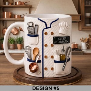 May include: A white ceramic mug designed to resemble a chef's coat, featuring a chef's hat, utensils, and the text "CHEF NORMAN." The mug has a large handle and is set on a wooden coaster. The design includes a grater, whisk, and small copper pan.