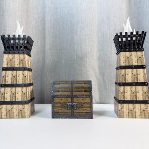 Könnte beinhalten: Ein dekoratives Set mit zwei holzartigen turmartigen Strukturen mit schwarzen Metallakzenten und einer kleinen Holzkiste. Die Türme haben eine käfigartige Oberseite mit weißen flammenförmigen Elementen. Die Truhe ist rechteckig mit Metalldetails.