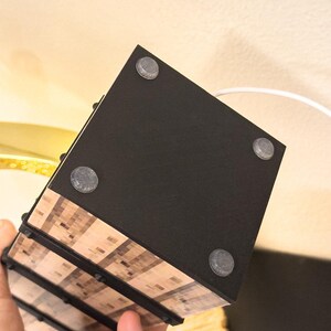 May include: A black, rectangular object with four gray, circular feet. The object has a wood-grain pattern on the sides and is held by a hand. A white cord extends from the top.