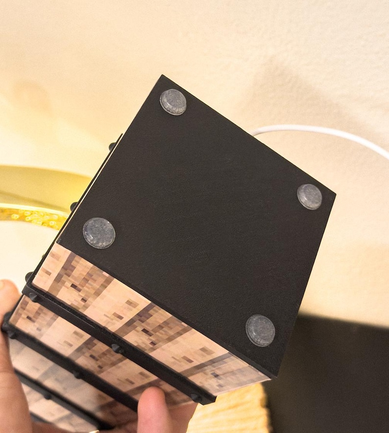 May include: A black, rectangular object with four gray, circular feet. The object has a wood-grain pattern on the sides and is held by a hand. A white cord extends from the top.
