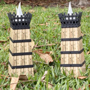 May include: Two decorative tower-shaped candle holders. The candle holders have a light brown, textured body with black metal bands and a black top. Each tower holds a white, flame-shaped candle. The candle holders are set in green grass.