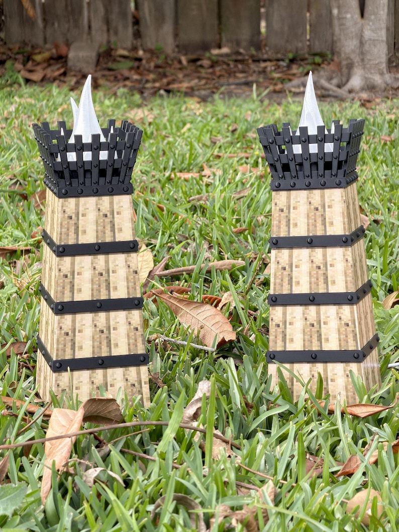 May include: Two decorative tower-shaped candle holders. The candle holders have a light brown, textured body with black metal bands and a black top. Each tower holds a white, flame-shaped candle. The candle holders are set in green grass.