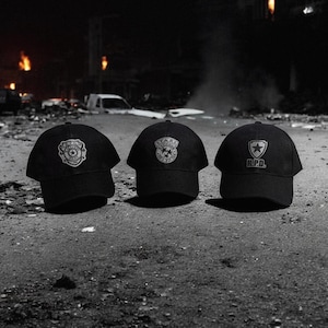 May include: Three black baseball caps with embroidered logos. The caps feature the emblems of R.P.D. and S.T.A.R.S. The caps are displayed on a gray surface, with a dark background suggesting a nighttime setting.
