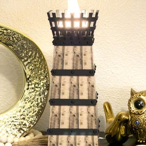 May include: A decorative tower-shaped lamp with a flickering flame effect. The lamp has a square base and a textured, pixelated design in shades of beige and brown. Black bands encircle the tower. The top is a black, castle-like structure.