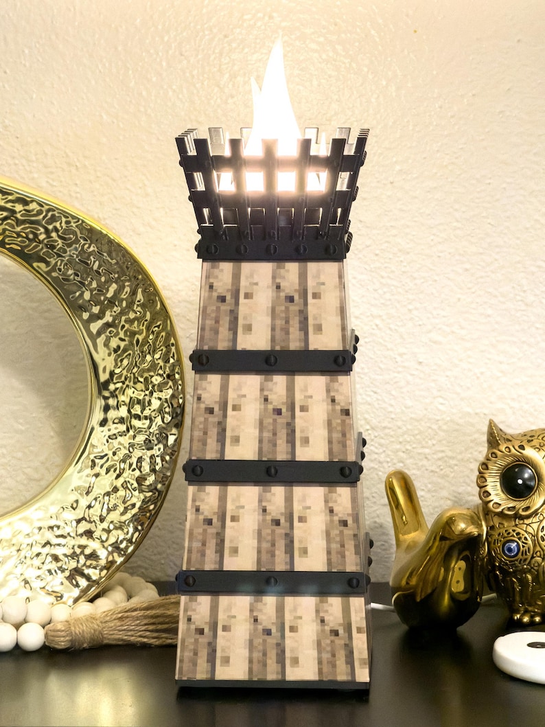 May include: A decorative tower-shaped lamp with a flickering flame effect. The lamp has a square base and a textured, pixelated design in shades of beige and brown. Black bands encircle the tower. The top is a black, castle-like structure.