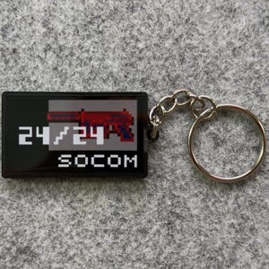 May include: Black rectangular keychain with a pixelated red and gray gun graphic. The text "24/24" and "SOCOM" are printed in white. The keychain has a silver key ring attached.