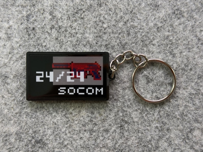 May include: Black rectangular keychain with a pixelated red and gray gun graphic. The text "24/24" and "SOCOM" are printed in white. The keychain has a silver key ring attached.