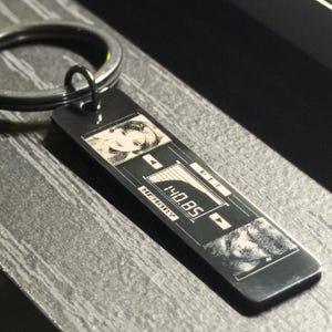 May include: A rectangular metal keychain with a silver finish. The keychain features a design with two grayscale images and the text "MEMORY" and "140.85". The keychain is attached to a silver key ring.