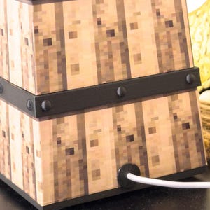 May include: A square lamp with a pixelated wood grain design. The lamp has black metal bands with rivets and a white power cord. The lamp is a decorative lighting fixture.