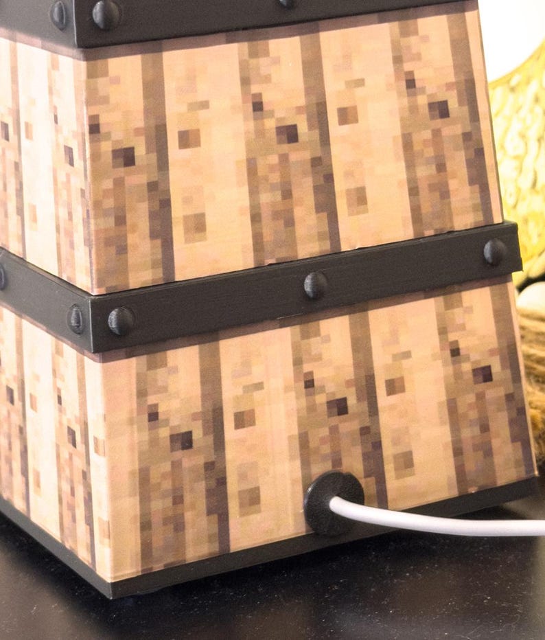 May include: A square lamp with a pixelated wood grain design. The lamp has black metal bands with rivets and a white power cord. The lamp is a decorative lighting fixture.