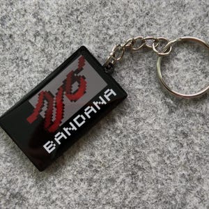 May include: Black rectangular keychain with a silver key ring and chain. The keychain has the word "BANDANA" in white and a red and white pixelated design on a gray background.