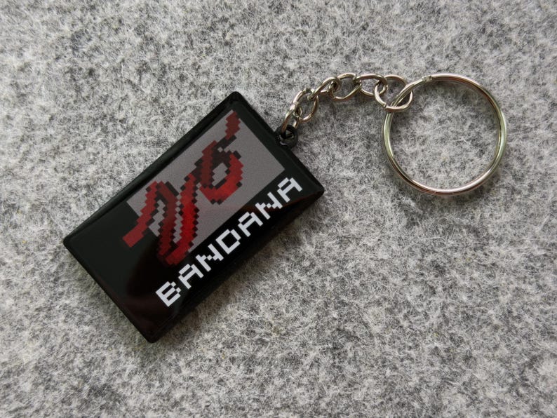 May include: Black rectangular keychain with a silver key ring and chain. The keychain has the word "BANDANA" in white and a red and white pixelated design on a gray background.