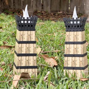 May include: Two decorative tower-shaped candle holders. The towers have a beige, vertical striped pattern with black metal bands and a black top with a white flame-shaped insert. They are set in a grassy area.