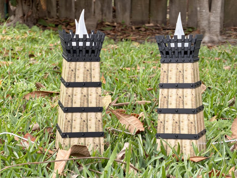May include: Two decorative tower-shaped candle holders. The towers have a beige, vertical striped pattern with black metal bands and a black top with a white flame-shaped insert. They are set in a grassy area.