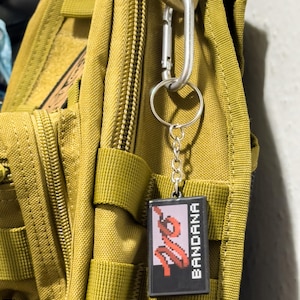 May include: A black keychain with the word "BANDANA" in white text and a red pixelated design. The keychain is attached to a silver carabiner clip, which is hooked onto a green backpack.