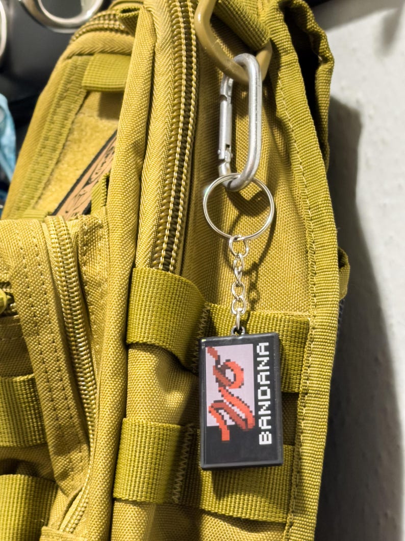 May include: A black keychain with the word "BANDANA" in white text and a red pixelated design. The keychain is attached to a silver carabiner clip, which is hooked onto a green backpack.