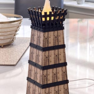 May include: A decorative lamp with a tower design. The lamp features a wooden-like exterior with black metal accents and a flickering flame-shaped light at the top. The lamp is powered by a white cord.