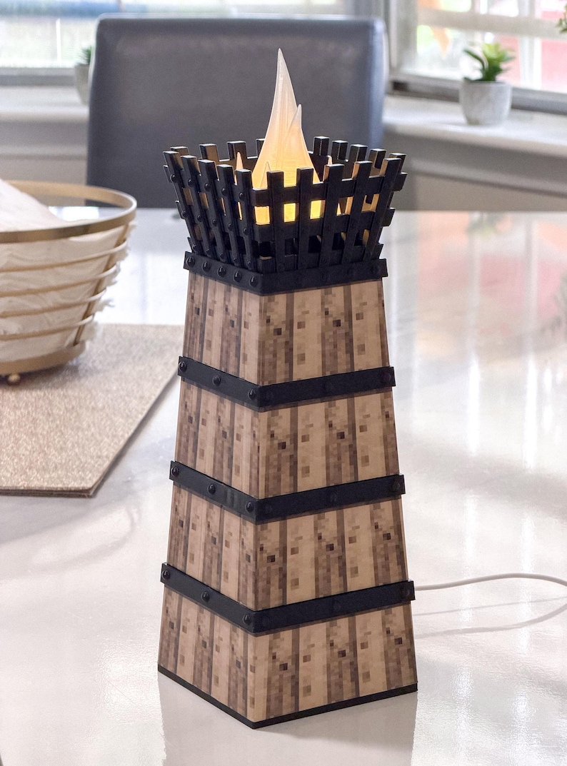May include: A decorative lamp with a tower design. The lamp features a wooden-like exterior with black metal accents and a flickering flame-shaped light at the top. The lamp is powered by a white cord.