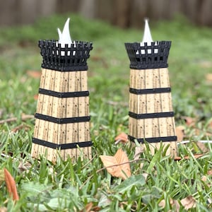 May include: Two decorative candle holders with a unique design. The holders are made of cork and black metal, with a cage-like top. The candles have a white flame-shaped light. The holders are placed on a bed of green grass.