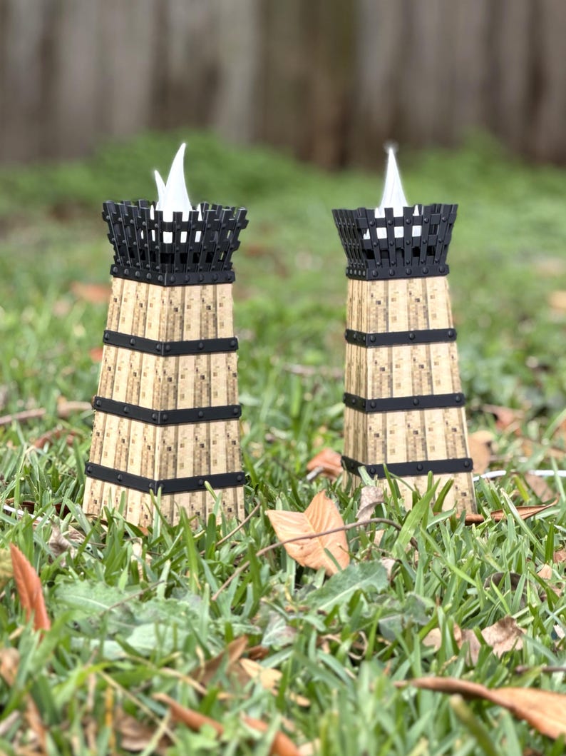 May include: Two decorative candle holders with a unique design. The holders are made of cork and black metal, with a cage-like top. The candles have a white flame-shaped light. The holders are placed on a bed of green grass.