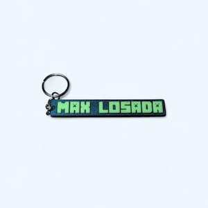 May include: A rectangular keychain with the text "MAX LOSADA" in green block letters. The keychain has a dark blue background with a glittery effect and a silver key ring.