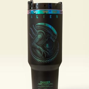 May include: Black tumbler with the word "ALIEN" in white and a detailed illustration of an alien creature. The tumbler has a black lid, a straw, and a holographic band around the top. The bottom of the tumbler has the word "DANGER" and other text.