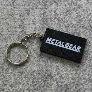 May include: Black rectangular keychain with the text "METAL GEAR" in white. The keychain has a silver-colored chain and a round key ring.