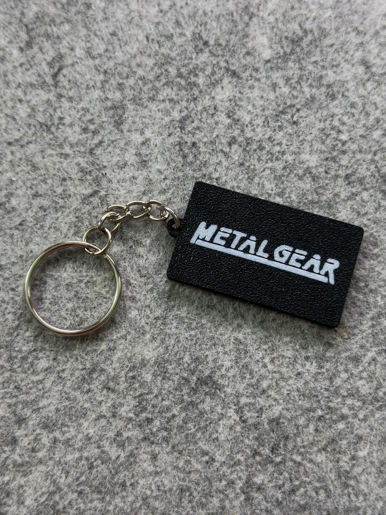 May include: Black rectangular keychain with the text "METAL GEAR" in white. The keychain has a silver-colored chain and a round key ring.